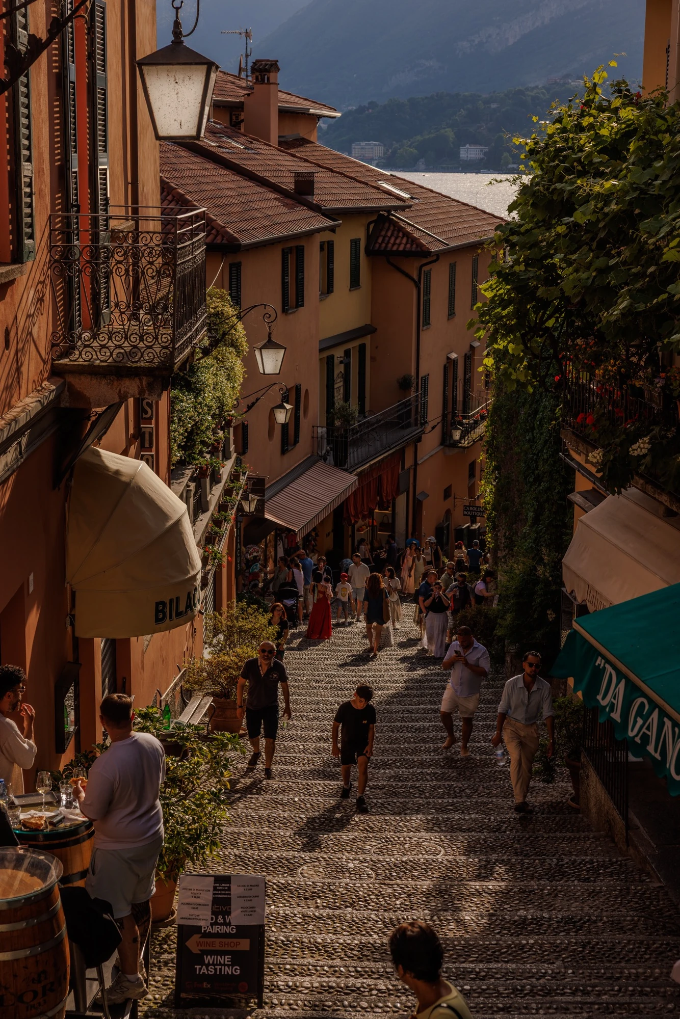 People walking down a narrow stepped street between colourful buildings above Lake Como.