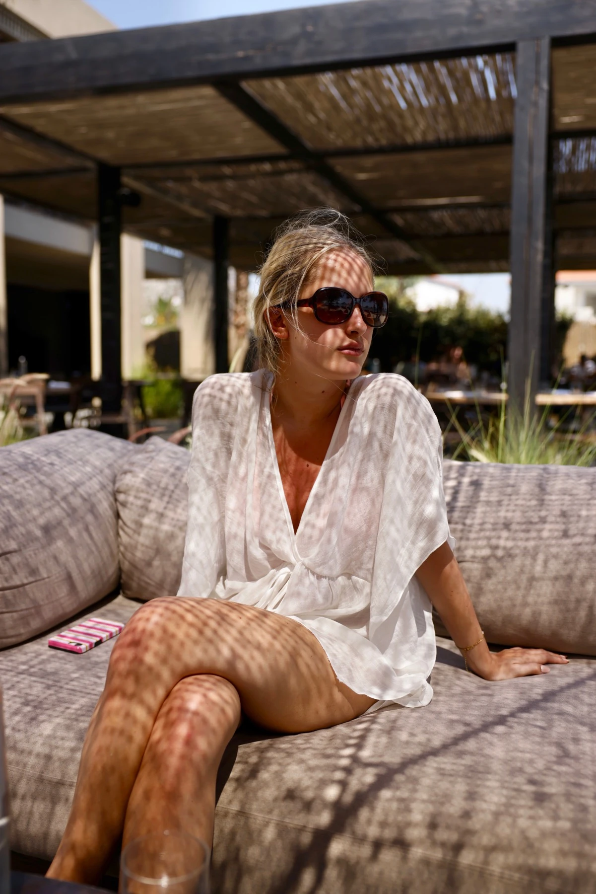 Woman wearing sunglasses and a white cover-up, seated on a sofa in patterned shade in Samos.